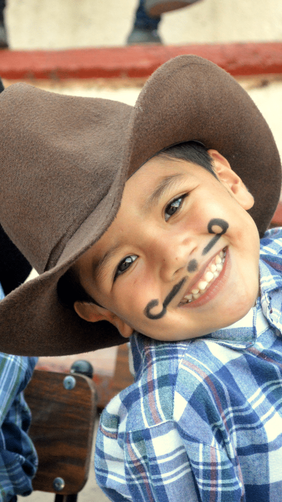 Junge mit einem aufgemaltem Bart, kariertem Hemd und einem Cowboyhut lächelt in die Kamera.